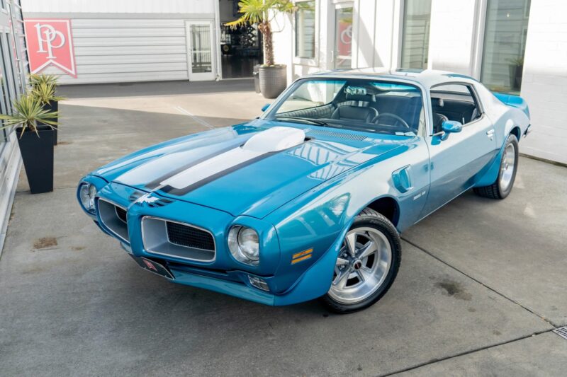 A blue classic Pontiac Firebird, a true icon among Classic Muscle Cars, with white racing stripes, parked on a concrete surface near a modern building with large windows.