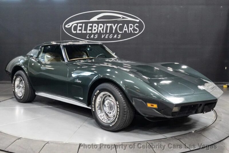 A dark green classic Chevrolet Corvette Stingray, a standout among Classic Muscle Cars, is displayed on a rotating platform in a showroom with a "Celebrity Cars Las Vegas" sign on the wall.