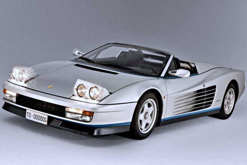 A silver convertible sports car—believed to be a 1-of-1: Rarest Ferraris In The World—features pop-up headlights, black side vents, and a front license plate reading “TO-000000G” on a neutral background.