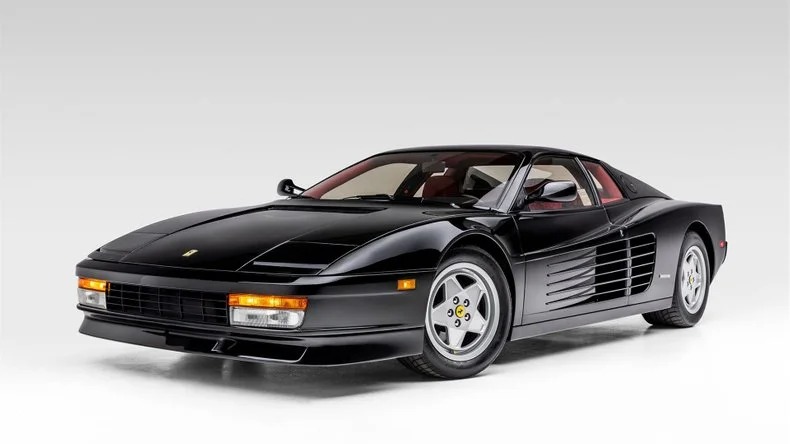 A black Ferrari Testarossa exotic car is parked in a studio setting with a neutral gray background.