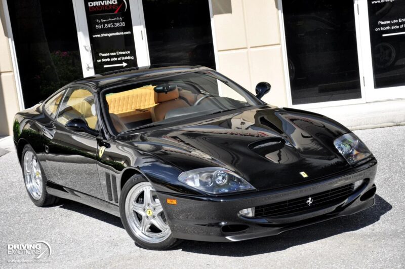 A black Ferrari 550 Maranello, a true luxury car for sale, is parked in front of a building with glass doors, its polished surface reflecting the sunlight.