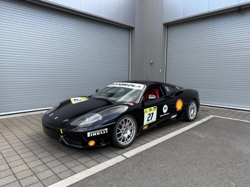 A black Ferrari luxury car with racing decals and sponsor logos is parked on a paved area in front of closed gray garage doors.
