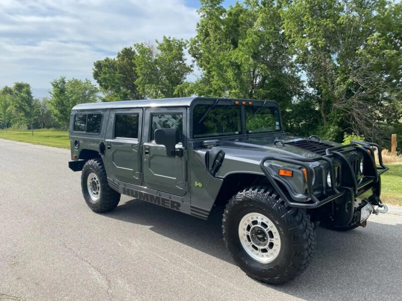 A black Hummer H1 luxury car SUV is parked on a paved road, surrounded by green trees and grass under a partly cloudy sky.