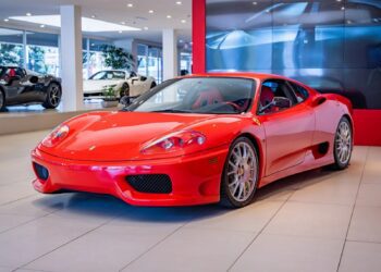 A red Ferrari luxury car is displayed in a brightly lit showroom, with other cars for sale visible in the background.