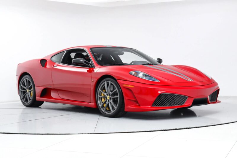 A 2000s Ferrari 430 Scuderia with black and silver stripes, yellow brake calipers, and a low-slung profile is parked in a white studio setting, for sale now.
