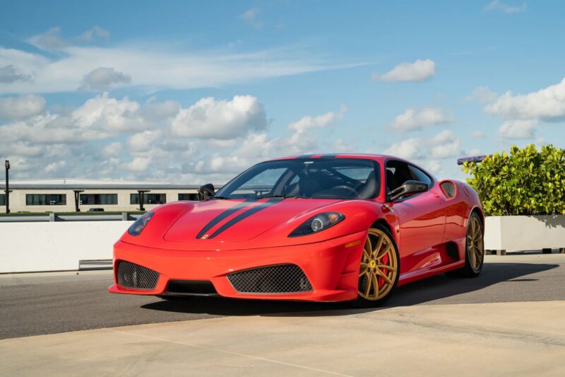A red Ferrari 430 Scuderia with black racing stripes and gold wheels is parked outdoors on a sunny day, with a building and blue sky in the background—a true 2000s Prancing Horse masterpiece.
