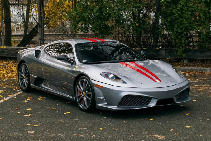 A silver Ferrari 430 Scuderia with red racing stripes is parked in an outdoor lot, surrounded by fallen leaves and trees with autumn foliage—a striking showcase of the Prancing Horse.