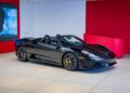 A black Ferrari 430 Scuderia convertible, showcasing the iconic Prancing Horse, is displayed in a modern showroom with white floors and red walls, ready for sale.