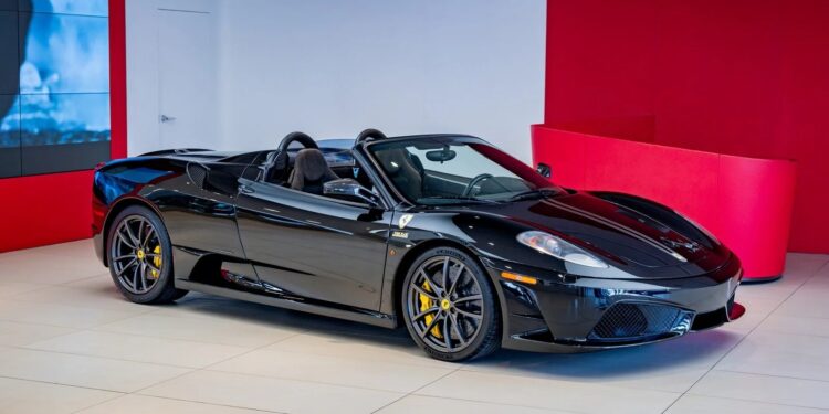 A black Ferrari 430 Scuderia convertible, showcasing the iconic Prancing Horse, is displayed in a modern showroom with white floors and red walls, ready for sale.