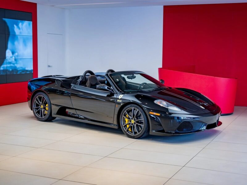A black Ferrari 430 Scuderia convertible, showcasing the iconic Prancing Horse, is displayed in a modern showroom with white floors and red walls, ready for sale.