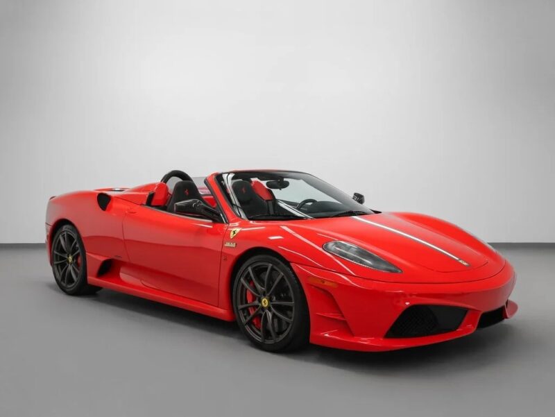 A Ferrari 430 Scuderia, sporting a prancing horse emblem, sits for sale indoors—a red convertible with black wheels and a gray racing stripe set against a plain white background.