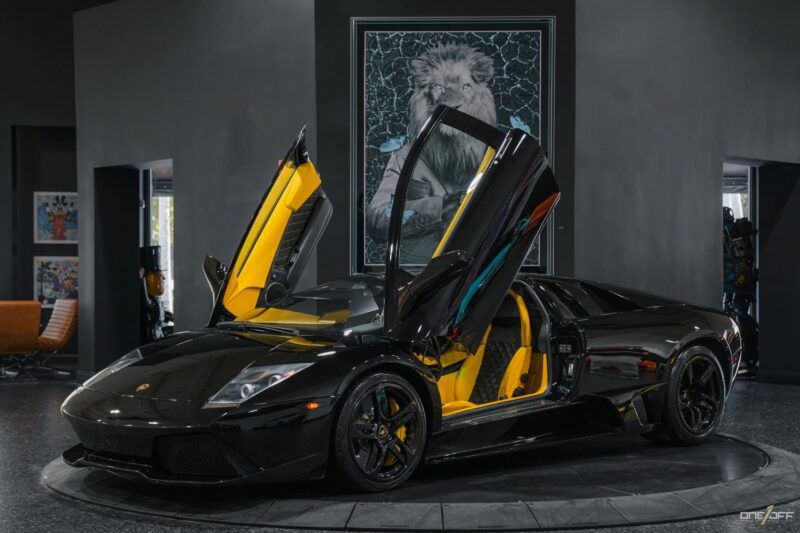 A black Lamborghini Murciélago exotic car with its scissor doors open is parked indoors; yellow interior accents are visible, and artwork hangs on the dark wall behind the car.