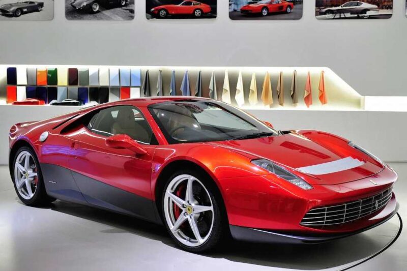 A red Ferrari sports car, one of the 1-of-1: Rarest Ferraris In The World, is on display in a showroom, with color and material samples on the wall behind it and framed car photos above.