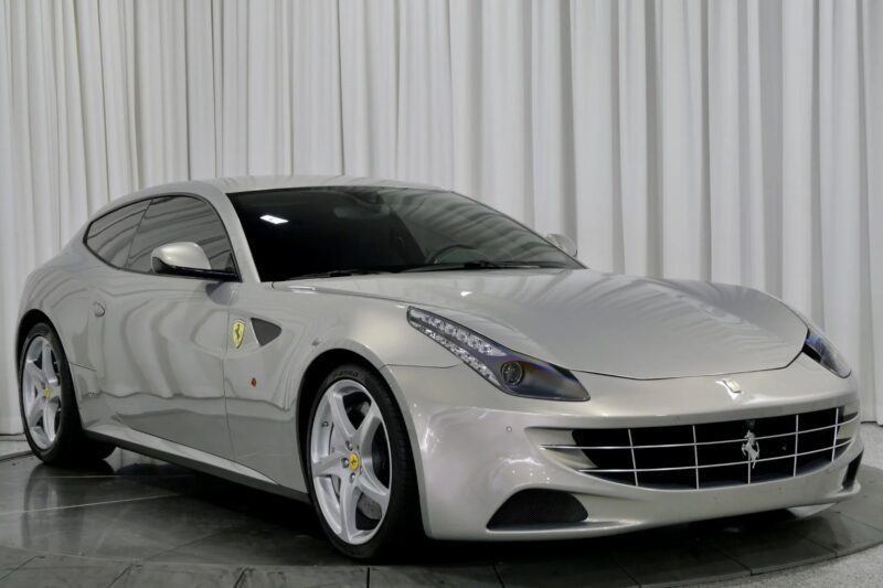 A silver Ferrari FF sports car, showcasing one of the elegant Ferrari colors, is parked indoors on a reflective floor with white curtains in the background.