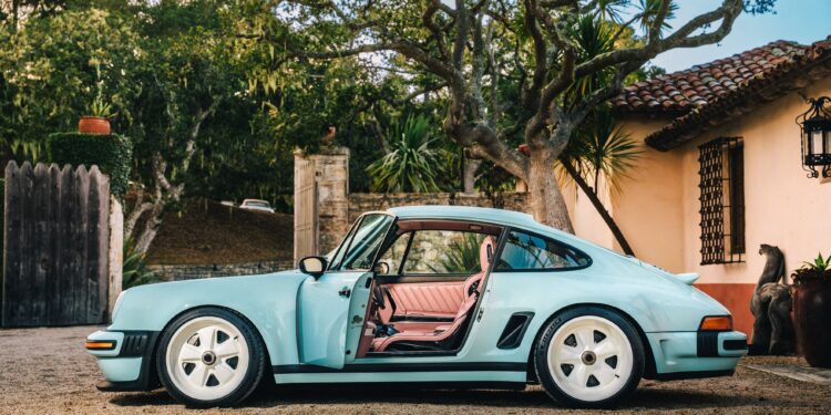 A light blue Reimagined Porsche 911 with white wheels is parked outside a house, the driver’s door open and trees in the background—a classic vision brought to life by Singer Vehicle Design.