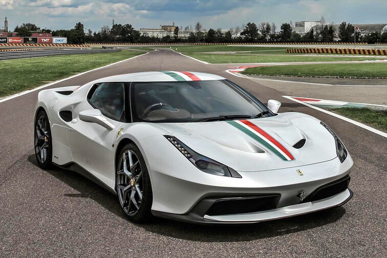 A white Ferrari 1-of-1, one of the rarest Ferraris in the world, with red and green stripes is parked on a race track with trees and buildings in the background.