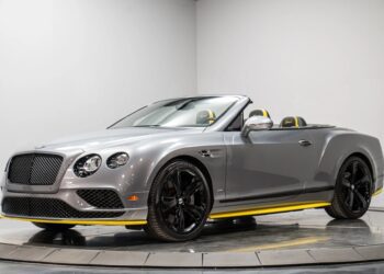 A silver Bentley Continental GTC convertible with black wheels and yellow accents is parked indoors on a rotating platform, embodying the essence of a refined luxury tourer.