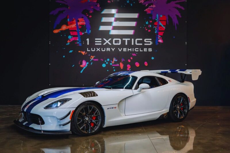 A white Dodge Viper luxury car with blue racing stripes is parked indoors in front of a black "1 Exotics Luxury Vehicles" sign with colorful palm tree graphics.