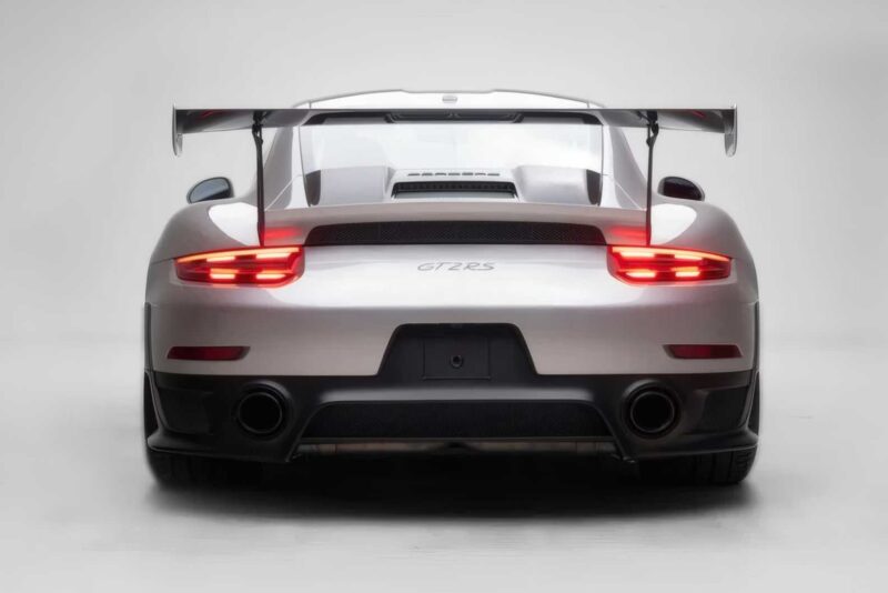 A rear view of a silver 2018 Porsche 911 GT2 RS, last of its kind before electrification, with illuminated taillights, a large rear wing, and dual exhaust pipes against a plain white background. For sale now.