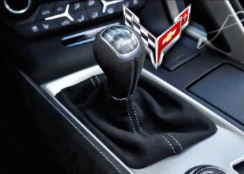 Close-up of a manual gear shift in a car, showcasing the Corvette logo near the lever—highlighting the craftsmanship of this Manual Production Corvette with dashboard controls visible in the background.