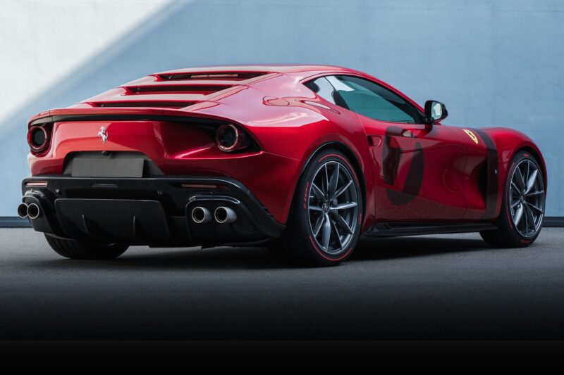 A red Ferrari sports car, one of the 1-of-1: Rarest Ferraris In The World, is parked on a smooth surface, shown from the rear side angle against a plain light blue wall.