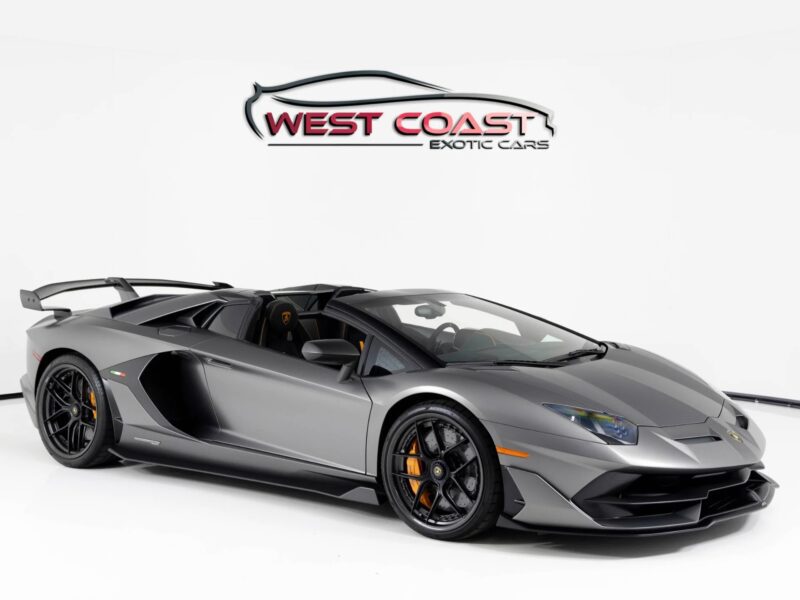 A matte grey Lamborghini Aventador convertible, a true luxury car, is displayed in a showroom with a "West Coast Exotic Cars" sign on the white wall behind it.