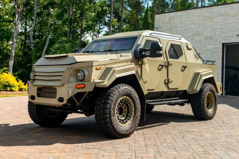 A beige, armored military-style vehicle with large off-road tires—resembling an exotic car—is parked on a paved driveway near a modern building and trees.