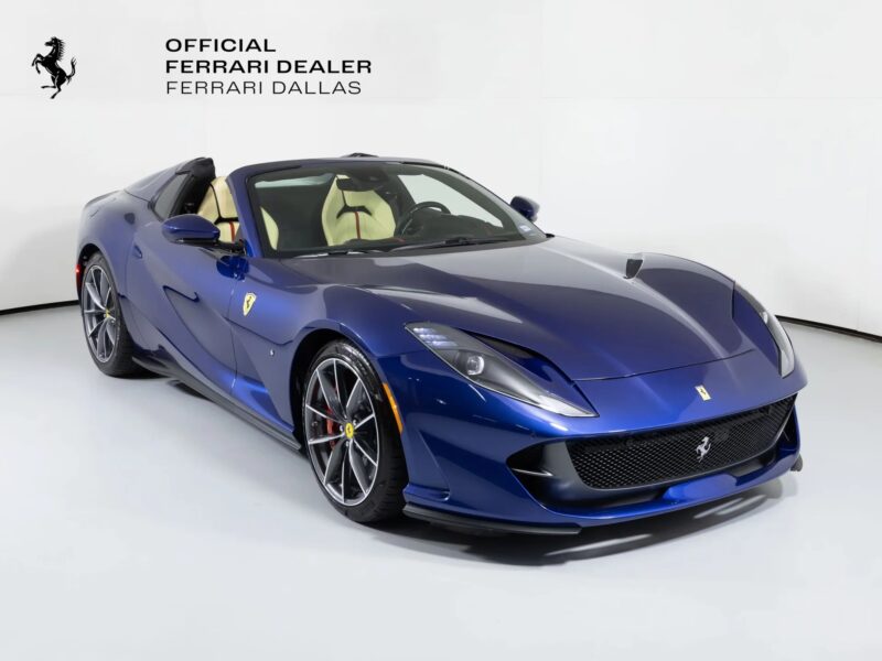 A blue Ferrari 812 GTS convertible in vibrant colors is displayed in a white showroom with "Official Ferrari Dealer Ferrari Dallas" signage on the wall, showcasing this stunning Ferrari for sale.