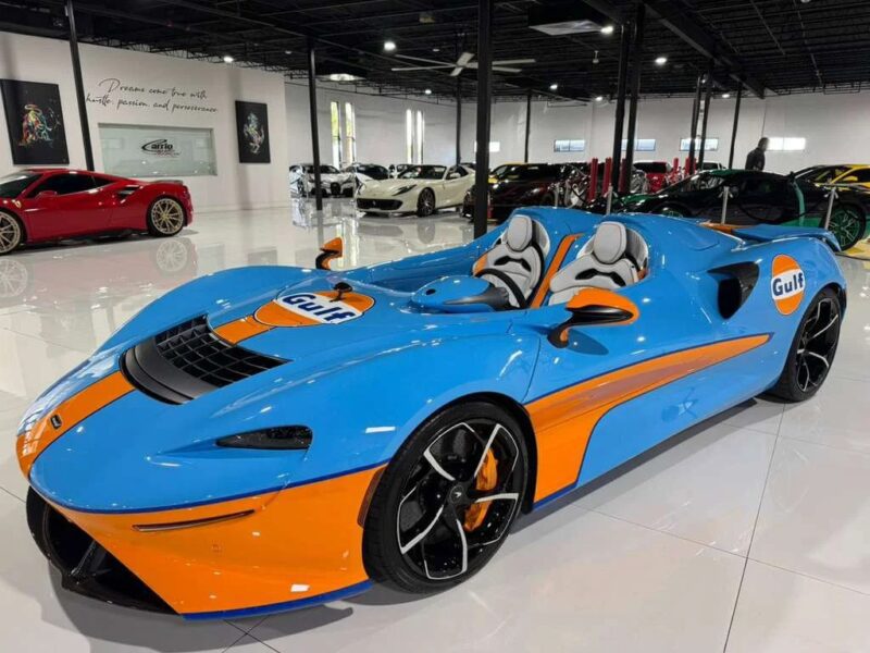 A blue and orange McLaren sports car with Gulf livery, an eye-catching exotic car, is displayed in a showroom surrounded by other luxury vehicles.