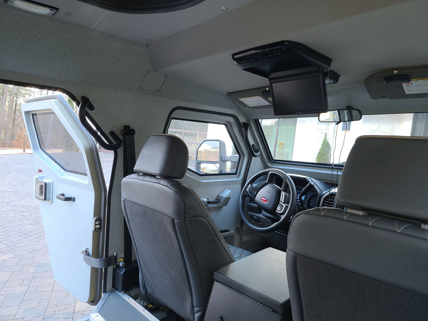 Interior view of a street-legal Terradyne Gurkha armored vehicle with driver and passenger seats, open rear door, and a ceiling-mounted monitor above the dashboard.