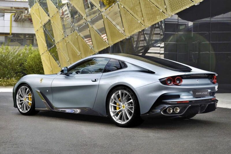 A silver Ferrari sports car, one of the 1-of-1: Rarest Ferraris In The World, is parked outdoors on a paved surface beside a modern building with a striking geometric gold-colored exterior feature.