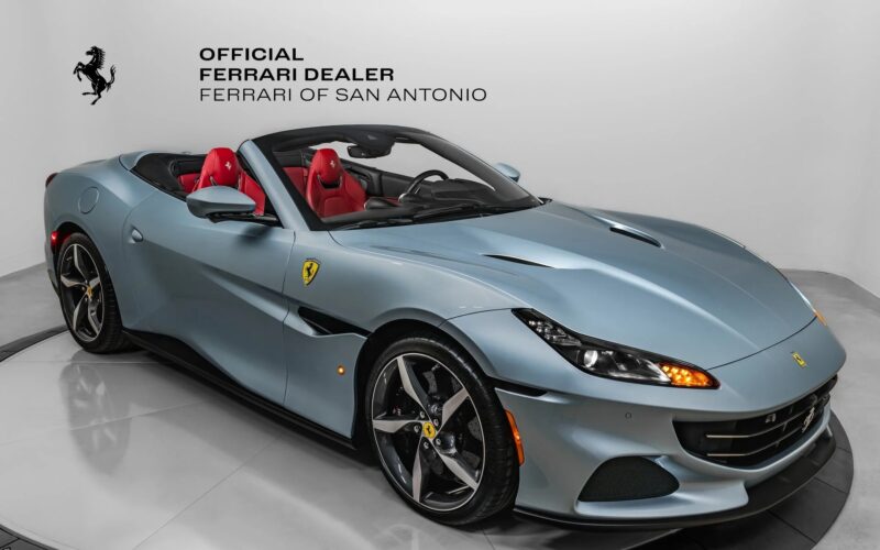 A metallic blue Ferrari convertible with a striking red interior is displayed in a showroom, showcasing iconic Ferrari colors beneath the "Official Ferrari Dealer Ferrari of San Antonio" logo on the wall.