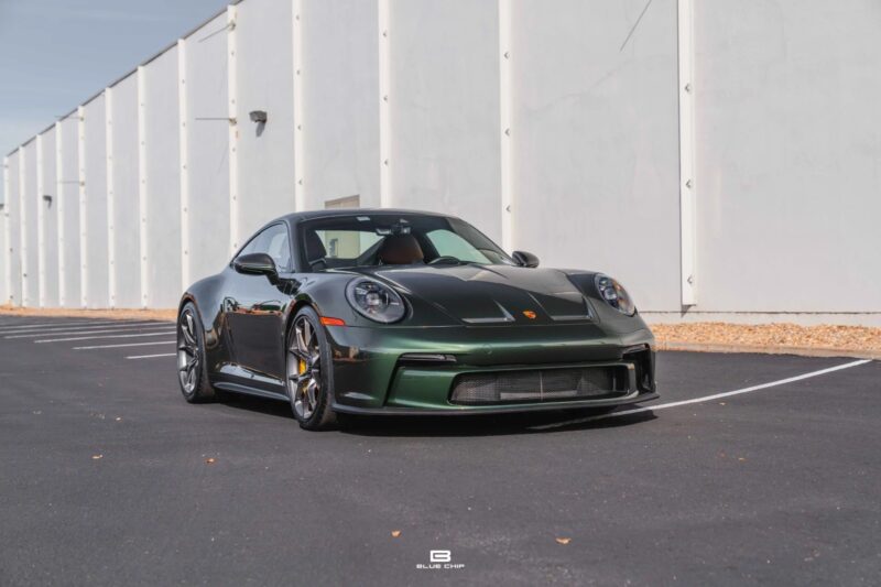 A dark green Porsche sports car, a true exotic car for sale, is parked in an empty lot next to a white industrial building under a clear sky.