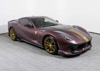 A maroon Ferrari 812 Competizione with gold racing stripes and V12 power, featuring gold wheels, is parked indoors on a gray tiled floor against a white wall.
