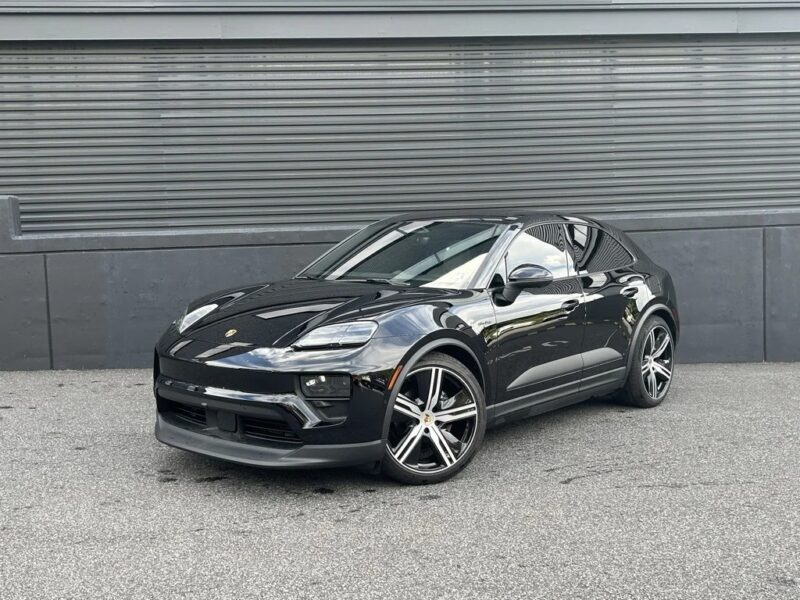 A black Porsche Macan EV luxury car is parked on a gray paved surface in front of a corrugated metal wall.