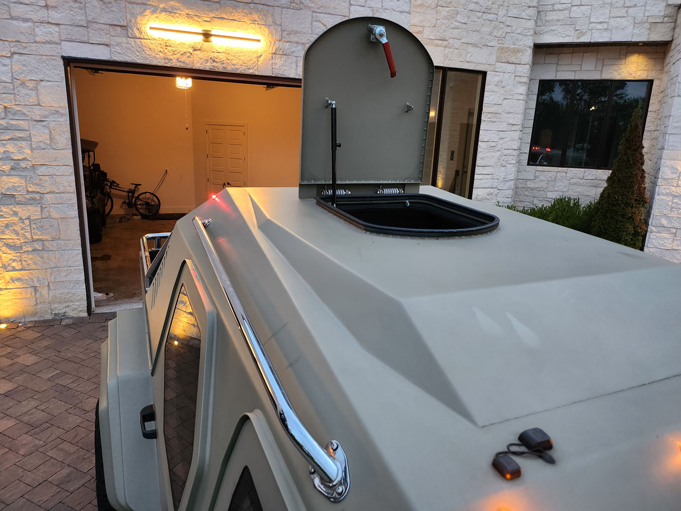 A street-legal Terradyne Gurkha military-style vehicle with an open roof hatch is parked outside a stone building with open garage doors and visible bicycles inside.