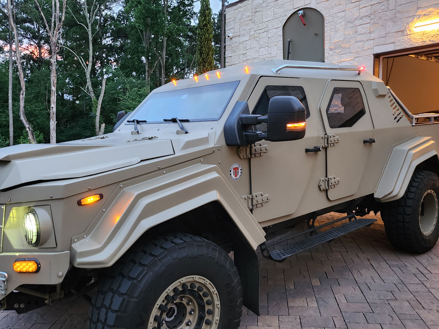 A beige, street-legal Terradyne Gurkha armored vehicle with large tires is parked on a brick driveway near a stone building and trees.