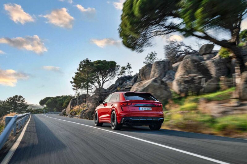 A red SUV, the 2025 Audi RS Q8 Performance, drives on a winding road bordered by rocks and trees under a partly cloudy sky. Review: Experience power and luxury on every curve.