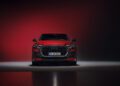 Front view of a red Audi SUV with headlights on, positioned against a dark red gradient background in a studio setting. Review: 2025 Audi RS Q8 Performance showcases bold design and dynamic presence in this striking image.