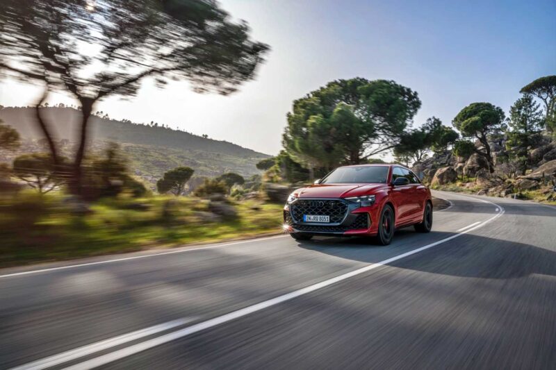 A red sports car, reminiscent of the Review: 2025 Audi RS Q8 Performance, drives along a curving, empty road surrounded by trees and hills under a clear sky.