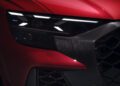 Close-up of the front left headlight and grille of a red car, featuring angular LED lights and a textured black grille design, as seen on the Review: 2025 Audi RS Q8 Performance.