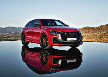 Review: 2025 Audi RS Q8 Performance—A striking red SUV parked on a reflective surface, set against a hilly landscape and clear sky.