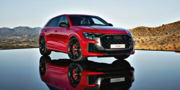 Review: 2025 Audi RS Q8 Performance—A striking red SUV parked on a reflective surface, set against a hilly landscape and clear sky.