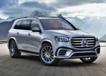 A silver Mercedes-Benz SUV, featured in the Review: 2025 Mercedes-Benz GLS 580 4Matic, is parked on a stone pavement with mountains and cloudy skies in the background.
