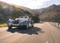 A classic Morgan Plus Four 75th Anniversary convertible drives along a winding road in a mountainous area, with wind turbines visible in the background.
