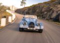 A light blue Morgan Plus Four vintage-style convertible car drives along a winding mountain road with rocky hills in the background, celebrating Morgan’s 75th Anniversary.