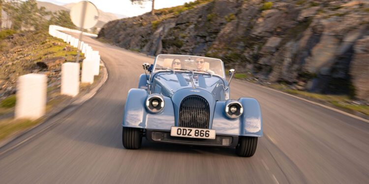 A light blue Morgan Plus Four vintage-style convertible car drives along a winding mountain road with rocky hills in the background, celebrating Morgan’s 75th Anniversary.