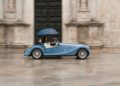 A person drives a blue Morgan Plus Four convertible in the rain, holding a blue umbrella and wearing white clothing, celebrating the car's 75th Anniversary.