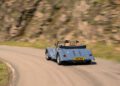 A light blue Morgan Plus Four vintage convertible with two people inside drives along a winding mountain road, surrounded by blurred rocky scenery.