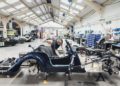 Two workers assemble classic Morgan cars, including a Morgan Plus Four 75th Anniversary edition, in a bright, spacious automotive workshop filled with tools, parts, and partially built vehicles.